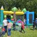 Eine große bunte Hüpfburg steht auf einer Wiese. Davor liegen viele Kinderschuhe und einige Erwachsene unterhalten sich.