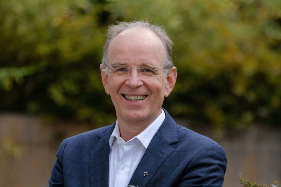 Ein Mann mit Halbglatze und blauem Anzug steht lächelnd im Grünen.