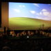 Blick in den Kinosaal: Auf der Leinwand eine grüne Wiese vor einem blauen Himmel. Vor der Leinwand sitzen Schafe und Hirten auf der Wiese.