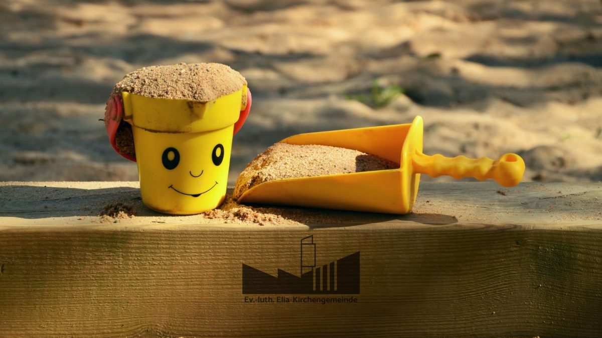 Ein kleiner Eimer und eine kleine Schaufel voll Sand liegen auf einem Holzbalken, der eine Sandkiste begrenzt.