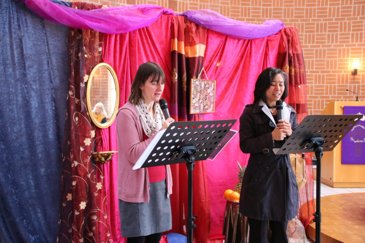 Zwei Frauen stehen vor einer orientalisch wirkenden Kulisse. Sie halten jeweils ein Mikrofon und haben einen Notenständer vor sich.