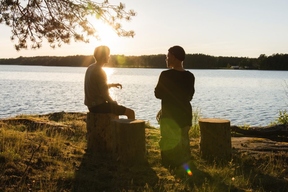 Zwei Männer sitzen auf Baumstümpfen an einem See, hinter dem die Sonne untergeht.