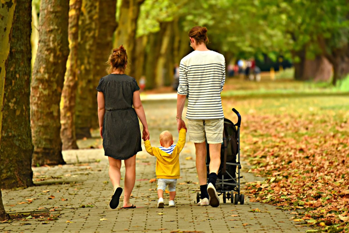 Eine Frau und ein Mann gehen auf einem Weg neben Bäumen. Ziwschen ihnen geht ein kleines Kind, dass sie an den Händen halten.