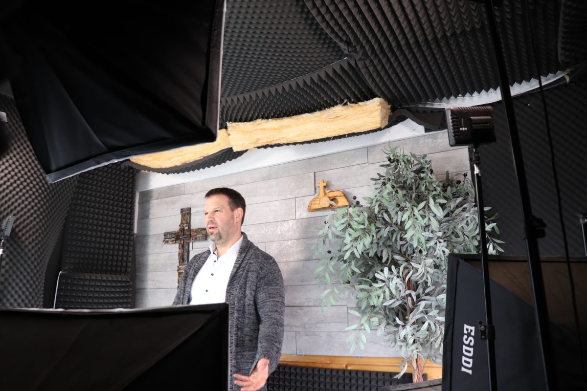 Blick in ein improvisiertes Film-Studio: mehrere Scheinwerfer und viele schallisolierende Elemente rahmen einen Mann in weißem Hemd und grauer Strickjacke ein, der vor einer hellgrauen Wand steht.